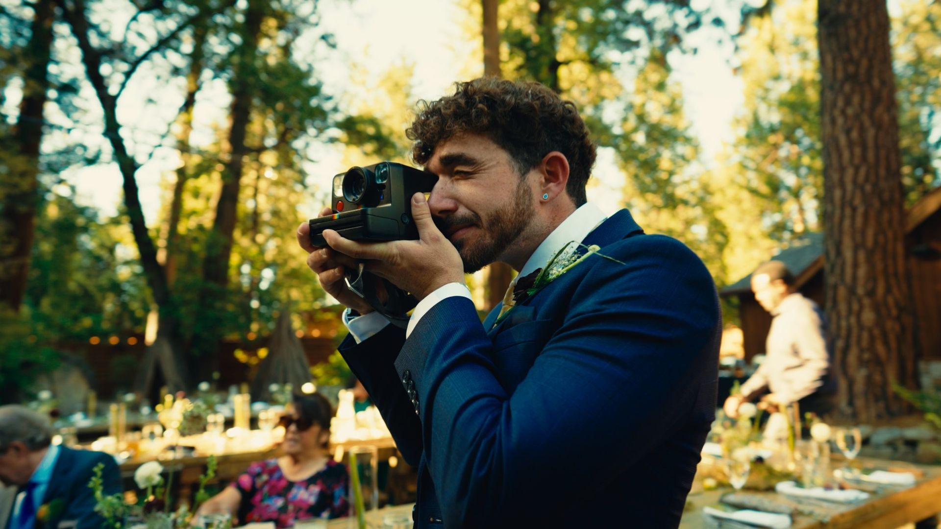 A wedding guest in a navy suit taking a Polaroid at a reception under tall pines