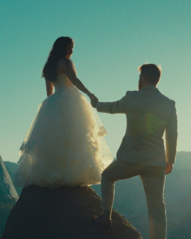 Groom helping his bride up onto a rock at Glacier Point, backlit by the sun